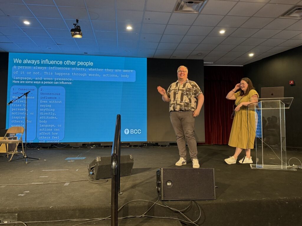 En mann står på scenen og ler, bak ham er presentasjon som foreteller om musikk